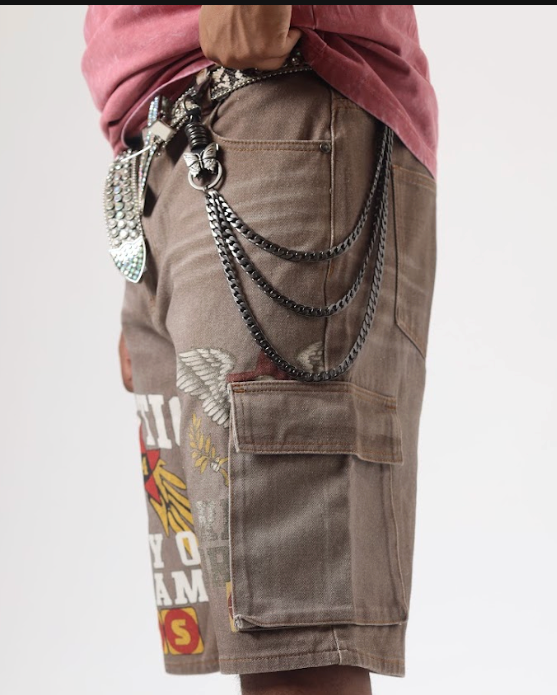A person shown from the waist down wearing brown cargo shorts with a graphic print and multiple hanging silver chains, paired with tan lace-up boots against a plain white studio background.